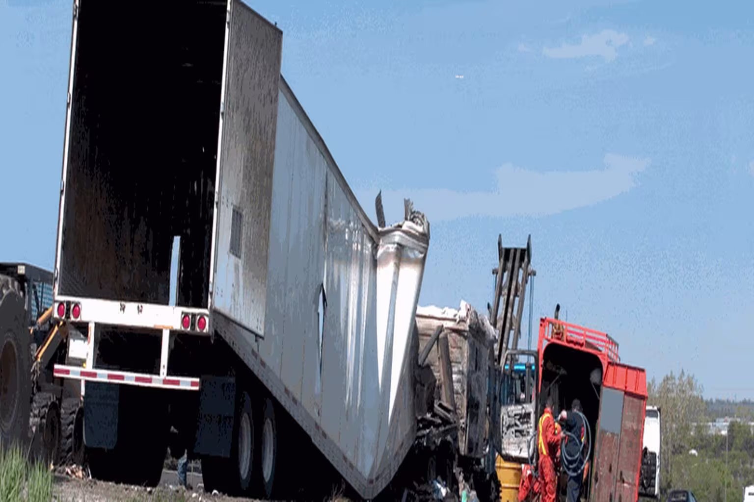 Damaged semi-truck trailer at an accident scene with workers and heavy equipment nearby.