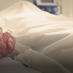 Hands holding each other over a bed with white sheets.