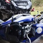 Close-up of a motorcycle accident scene showing a blue motorcycle lying on its side in front of a heavily damaged black car