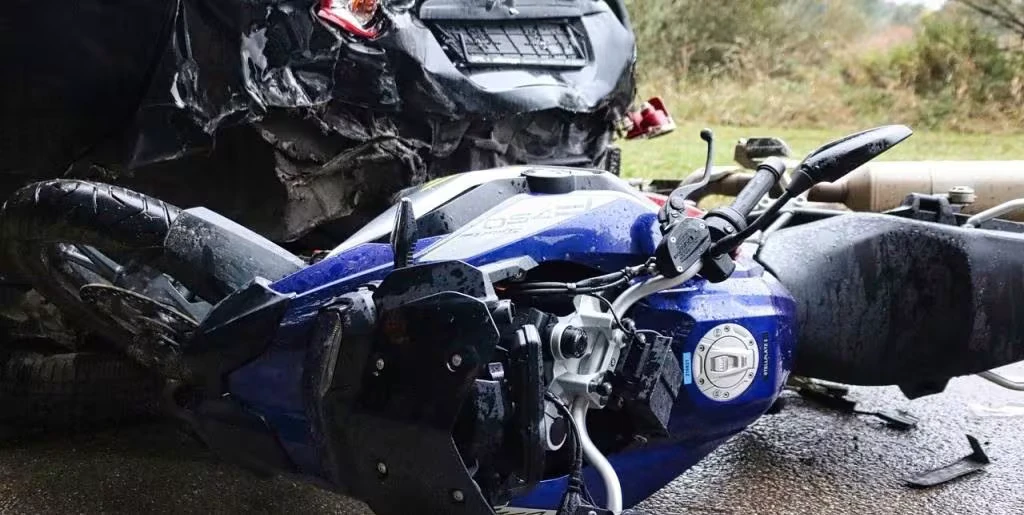 Close-up of a motorcycle accident scene showing a blue motorcycle lying on its side in front of a heavily damaged black car