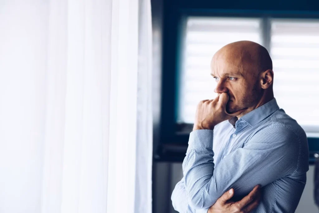 Worried man in business attire looks out a bright window, reflecting on a difficult situation.