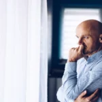 Worried man in business attire looks out a bright window, reflecting on a difficult situation.