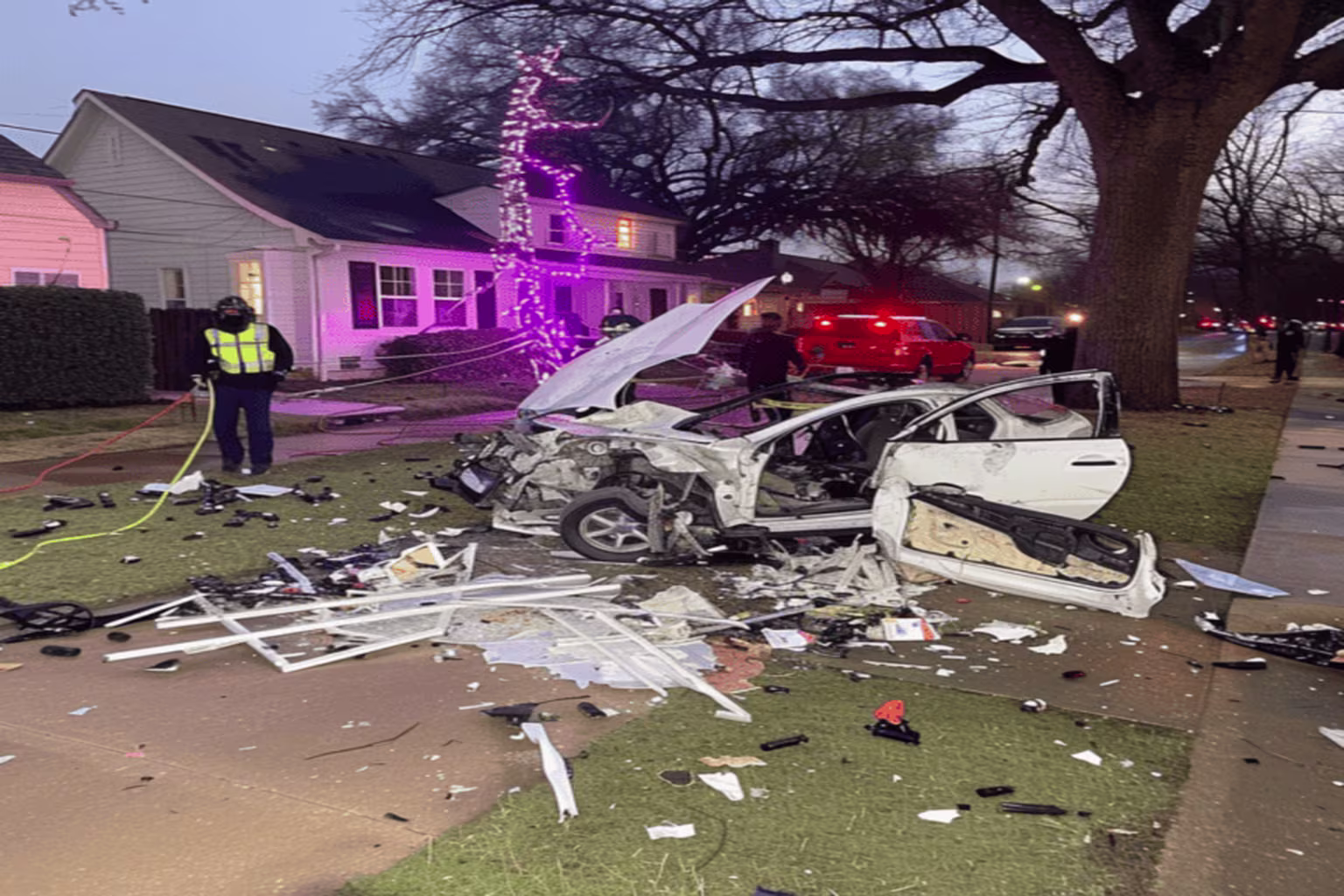 Car crash scene with a damaged vehicle on the lawn and emergency responders present.