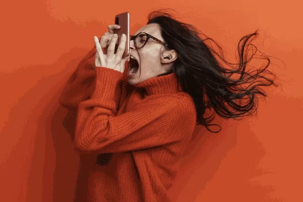 Frustrated woman in an orange sweater yelling into her phone against an orange background.