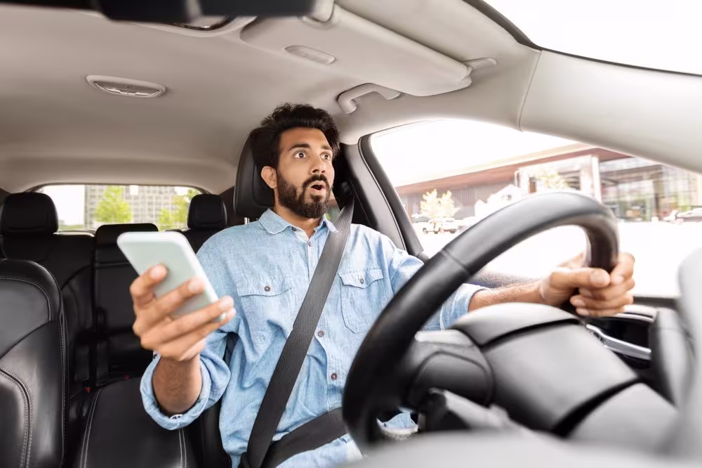 Surprised driver in blue shirt holding phone while driving, looking shocked with one hand on steering wheel