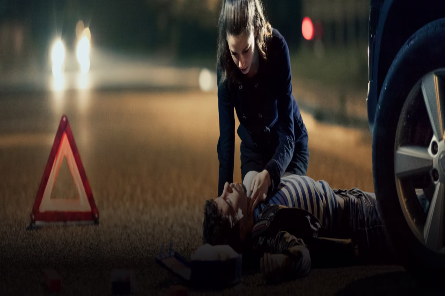 A person assists an injured individual on a road at night.