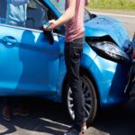 Two drivers inspect damage after a rear-end collision between a blue car and a gray car.