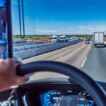 View from truck driver's seat showing hands on steering wheel while driving on busy Texas highway with multiple commercial trucks, illustrating hours of service regulations and road safety