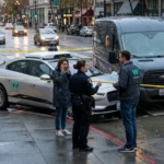 Self-driving car and van involved in a crash at a city intersection, with police officers speaking to people as the scene is taped off on a rainy day.