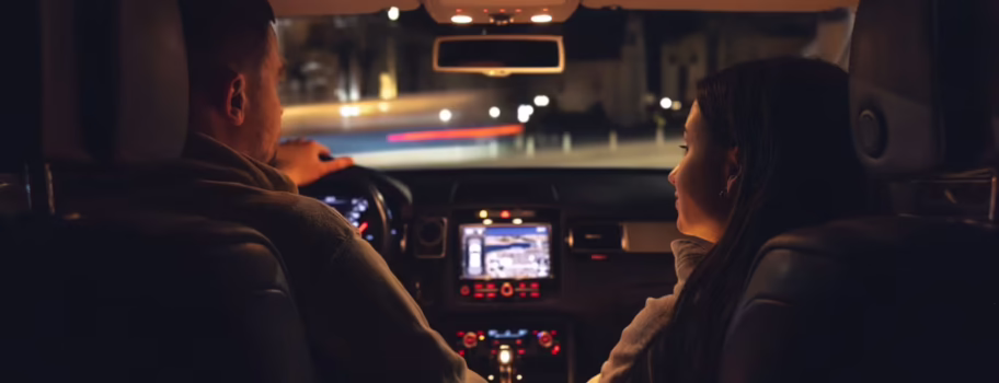 Two people driving at night with interior car lights on, illuminating the cabin while city lights are visible outside
